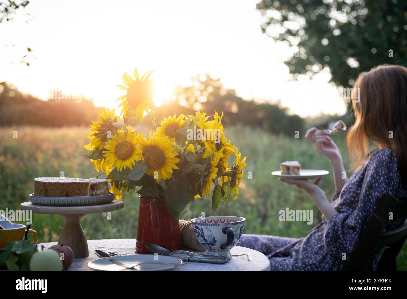 una niña se sienta en el jardín y come un pastel, un jarrón con girasoles y manzanas sobre una mesa Foto de stock