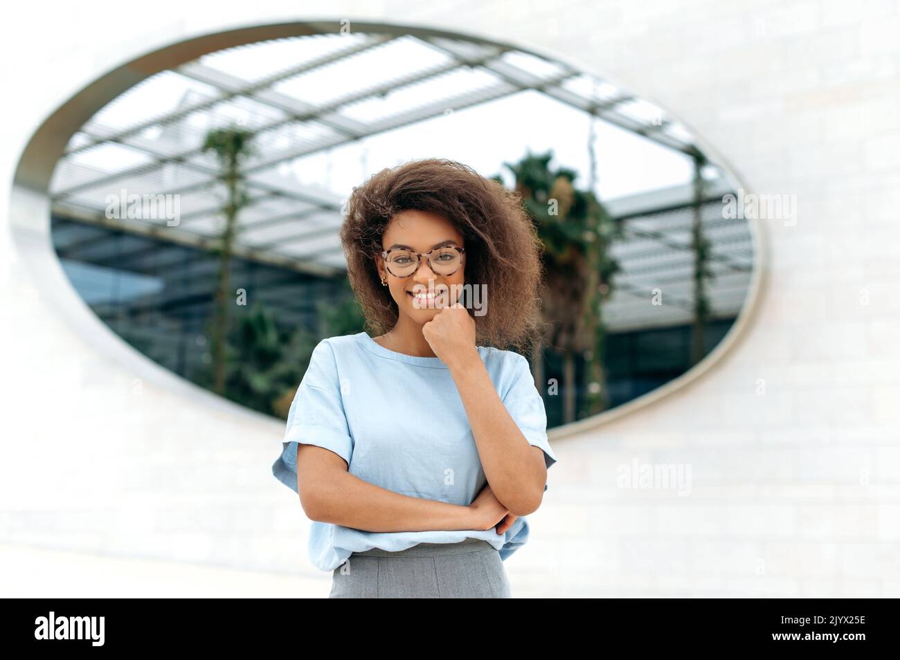 Retrato de una hermosa mujer afroamericana segura y positiva con pelo rizado, luciendo una camisa azul, con gafas, de pie al aire libre, mirando la cámara, sonriendo amable Foto de stock