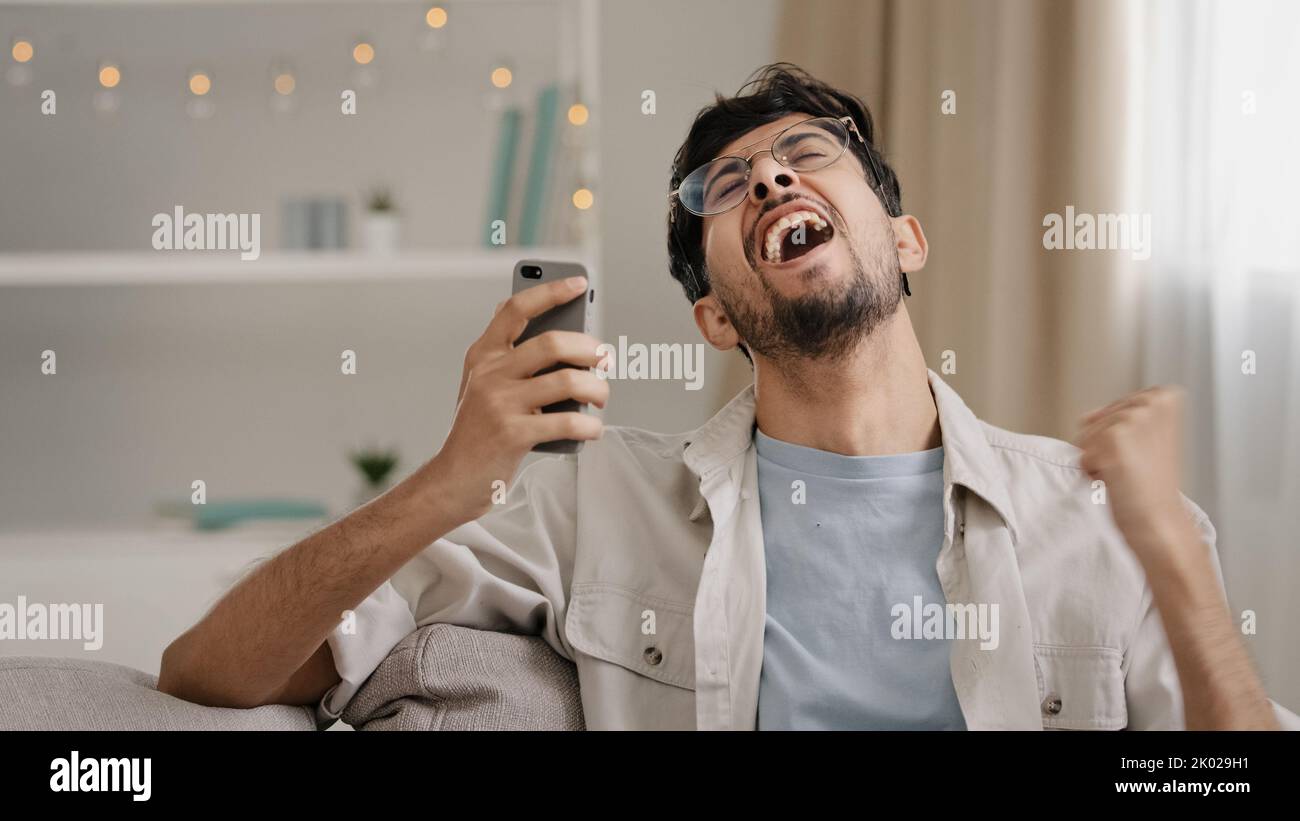 Un joven hombre barbudo árabe de 30s años con gafas se sienta en casa en el sofá mirando el teléfono móvil haciendo un gesto sí obtener oferta ganar buenas noticias Foto de stock