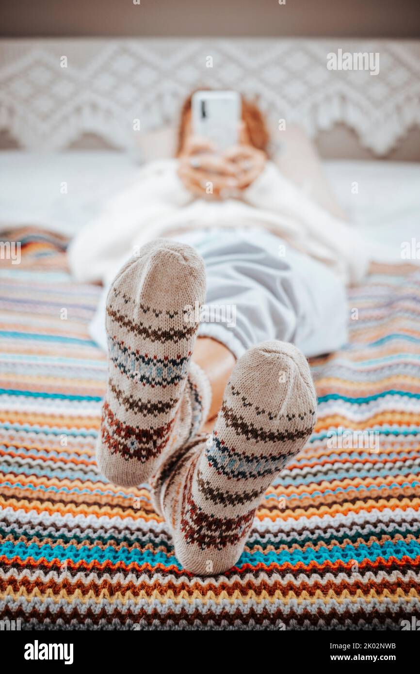 Primer plano de los pies y calcetines agradables en una misma textura ganchillo cobija de colores. Mujer que tiene relajarse actividad de ocio en el dormitorio y utilizar el teléfono móvil en el fondo desenfocado. Foto de stock