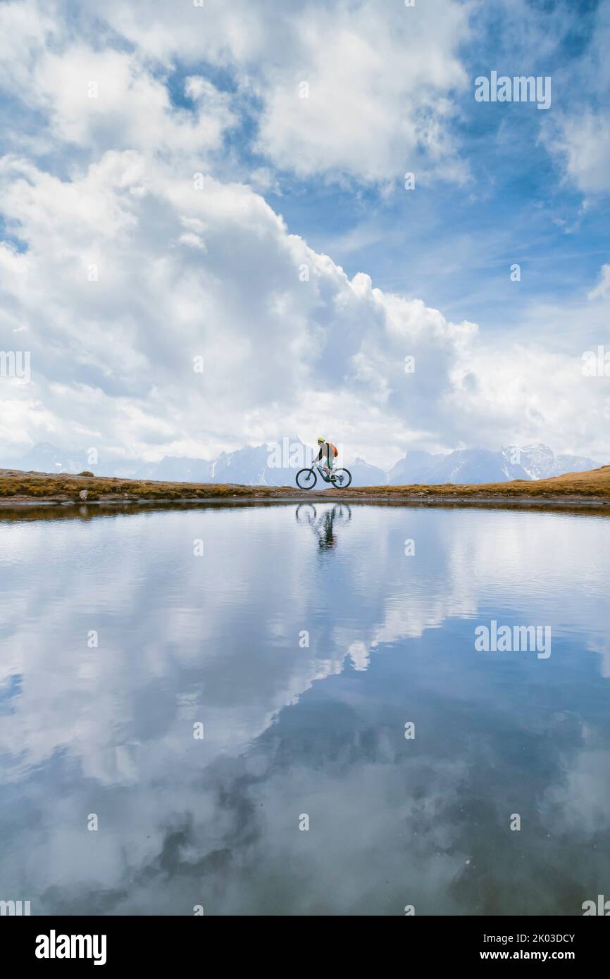 Italia, Tirol del Sur, Bolzano / Bozen, San Candido / Innichen. Ciclista con bicicleta electrónica, reflejo en un pequeño lago alpino en un día nublado Foto de stock