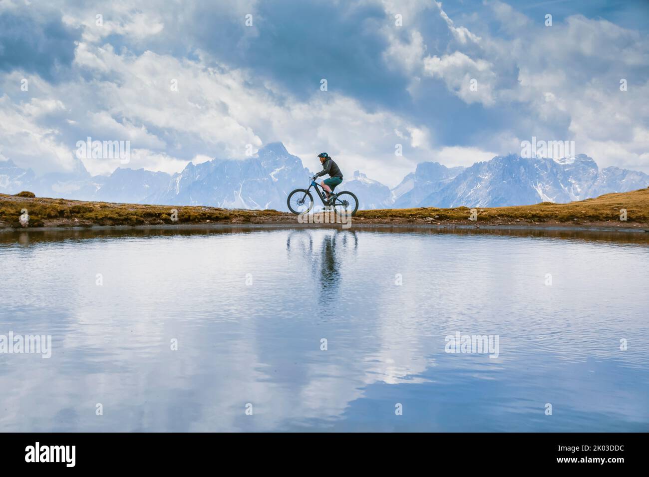 Italia, Tirol del Sur, Bolzano / Bozen, San Candido / Innichen. Ciclista con bicicleta electrónica, reflejo en un pequeño lago alpino en un día nublado Foto de stock