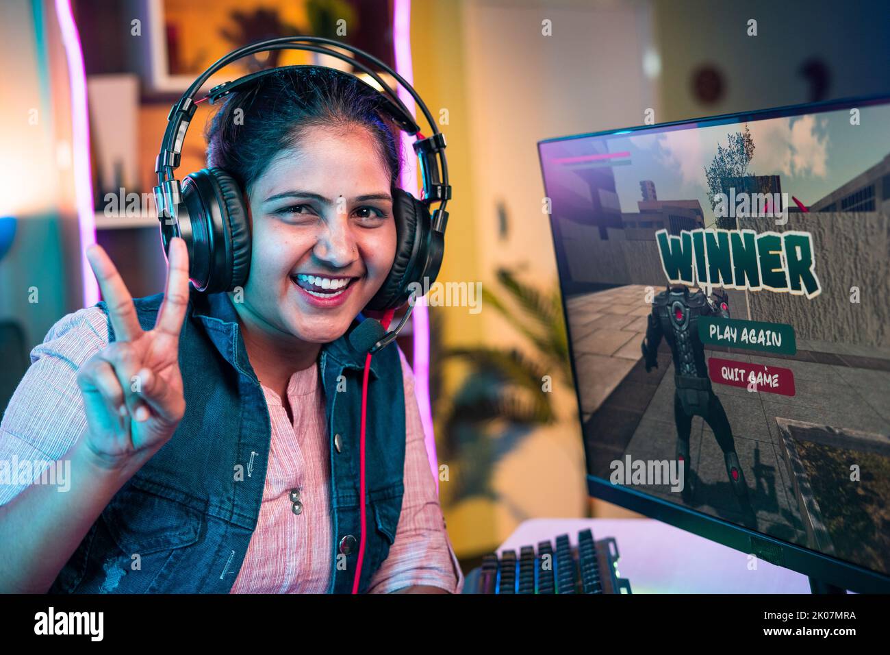 Sonriendo jugador con auriculares celebrando el juego de ganar mostrando signo de victoria mientras juega a videojuegos en vivo en el ordenador en casa - concepto de campeonato Foto de stock