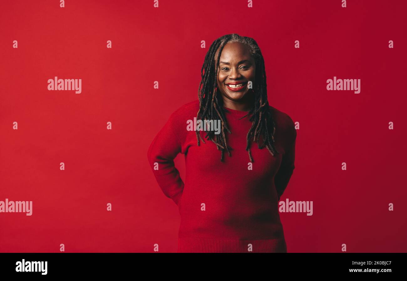 Mujer negra con los dreadlocks sonriendo en la cámara mientras que parado contra un fondo rojo. Mujer madura feliz abrazando su pelo natural con confiden Foto de stock