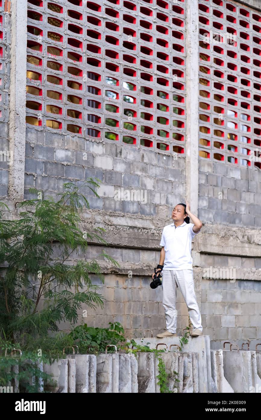 El camarógrafo profesional asiático es pretencioso y está en la construcción, en una barricada de hormigón y mira al cielo, y piensa en cómo publicar una fotografía Foto de stock