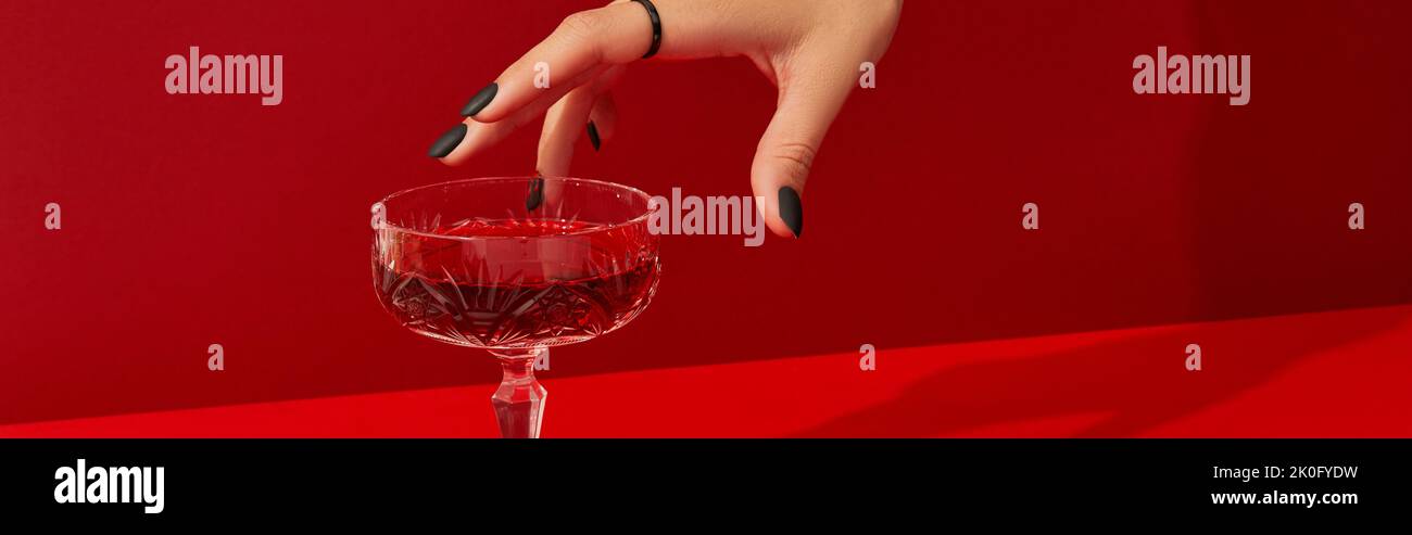 Mano de mujer con diseño de uñas espeluznante Tome vidrio con cóctel de halloween de arándanos sobre fondo rojo Foto de stock