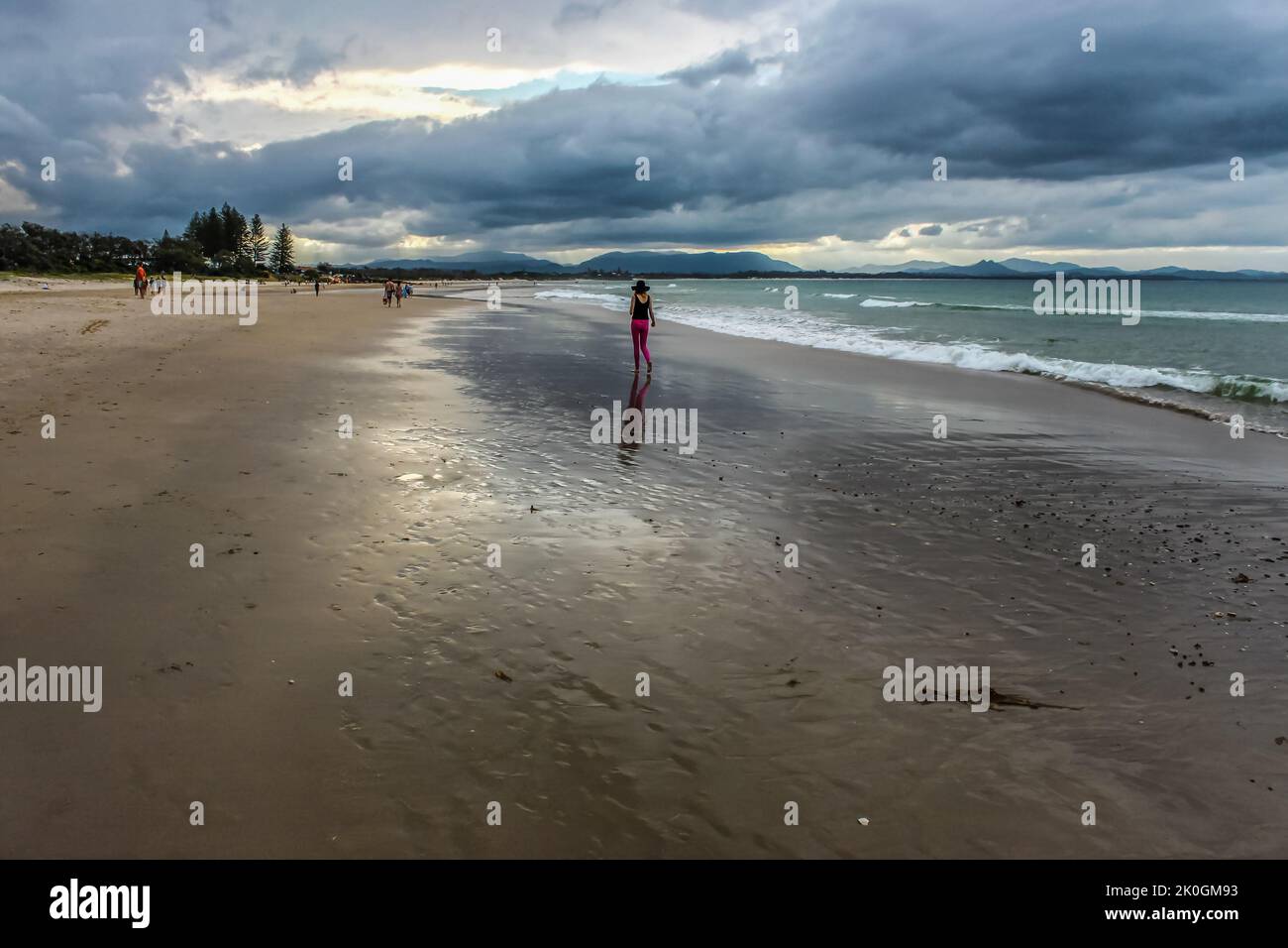 Mujer con leggings rosados y sombrero de ala ancha caminando por la playa húmeda bajo cielos tormentosos en Byron Bay Australia con otros amantes de la playa y montañas i Foto de stock