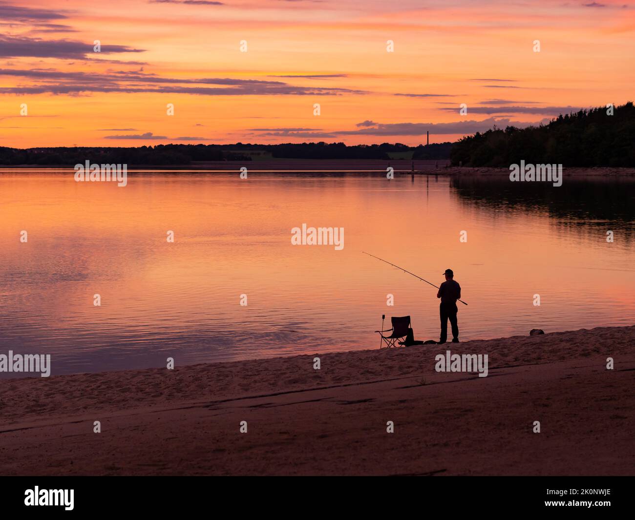Silueta de un pescador pescando en un lago durante la puesta del sol. Un pescador al anochecer con equipo como una silla de camping. La playa de arena es visible. Foto de stock
