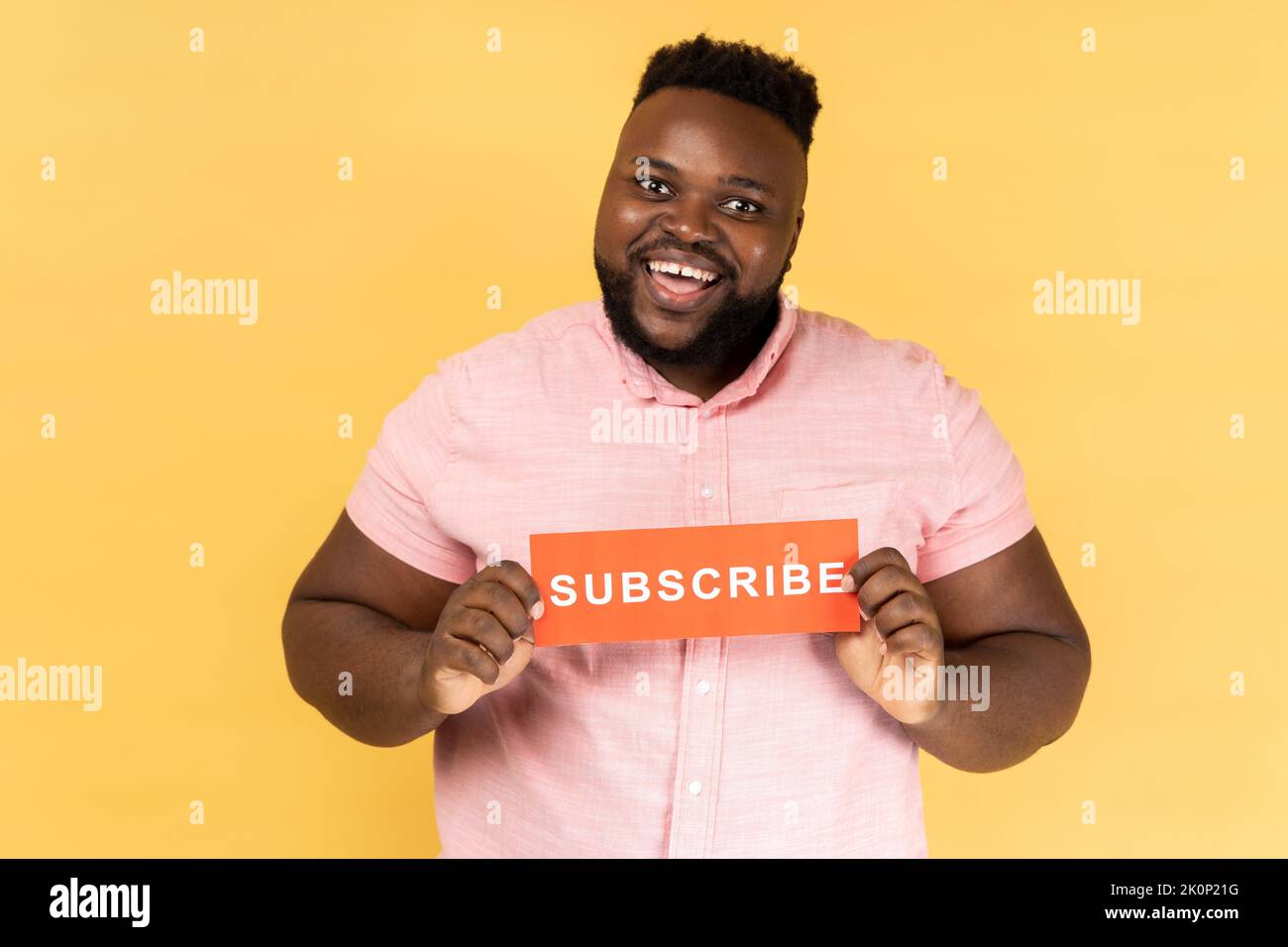 Retrato de alegre hombre barbudo positivo con camisa rosa sosteniendo tarjeta roja con inscripción suscrita, pidiendo seguir su vlog. Estudio de interior grabado aislado sobre fondo amarillo. Foto de stock
