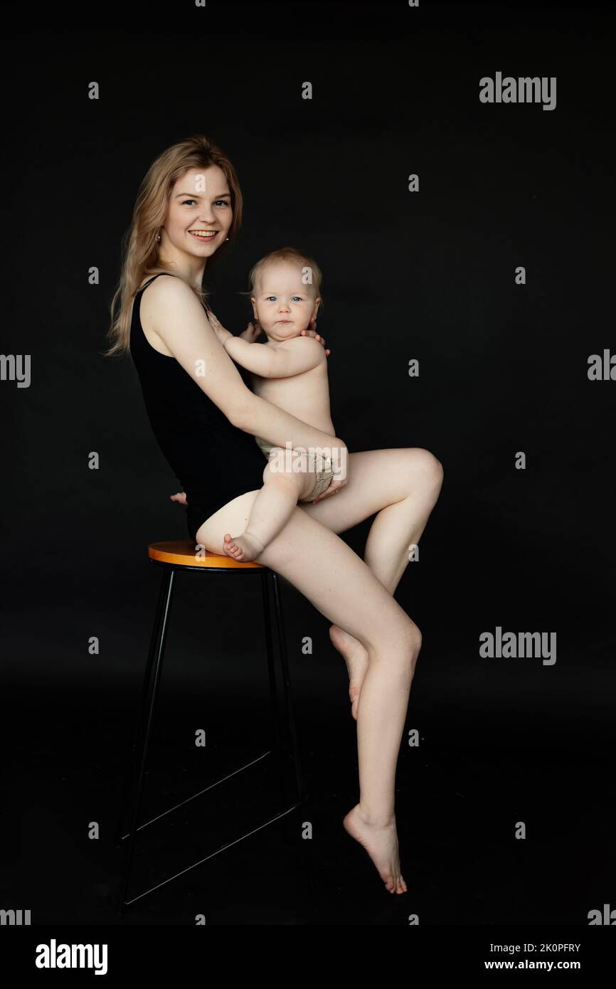 Retrato vertical de una joven mujer rubia y sonriente mirando la cámara sosteniendo al pequeño bebé, sentada en la silla sobre fondo negro Foto de stock