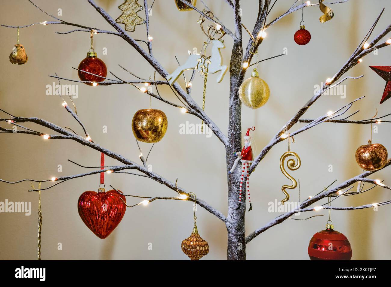 Adornos para el árbol de Navidad en rojo tradicional y dorado colgando en un moderno árbol de pelucas con efecto de nieve Foto de stock