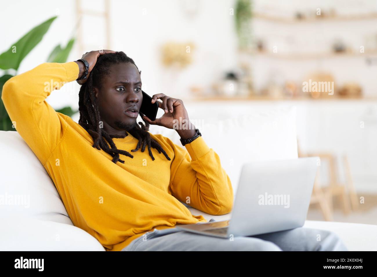 Estafa de Internet. Hombre negro sorprendido mirando el ordenador portátil y hablando por teléfono móvil Foto de stock
