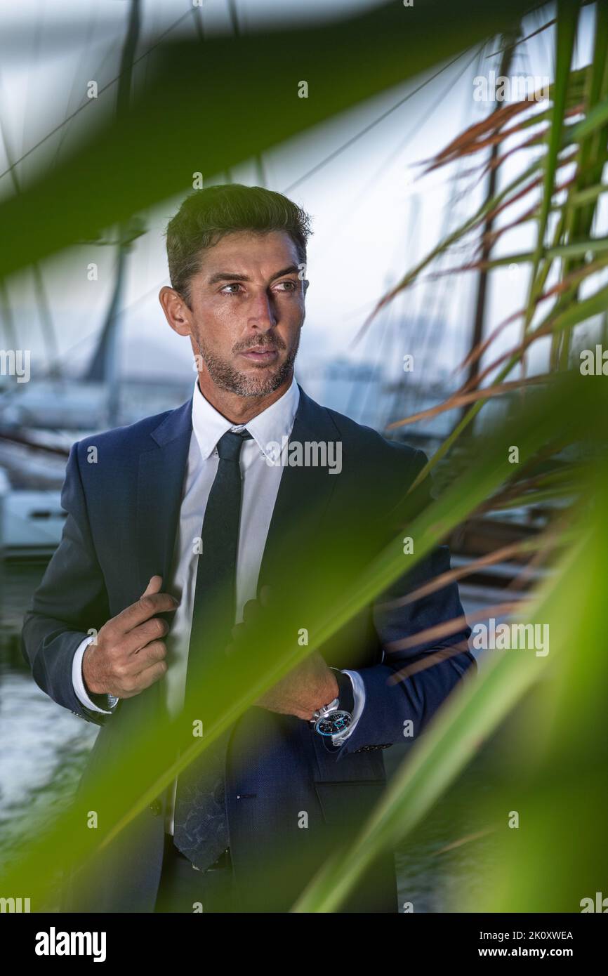 Hombre ejecutivo autoasegurado en traje chaqueta de vestir y mirar a distancia mientras se encuentra detrás de las hojas de palma en el puerto de la ciudad Foto de stock