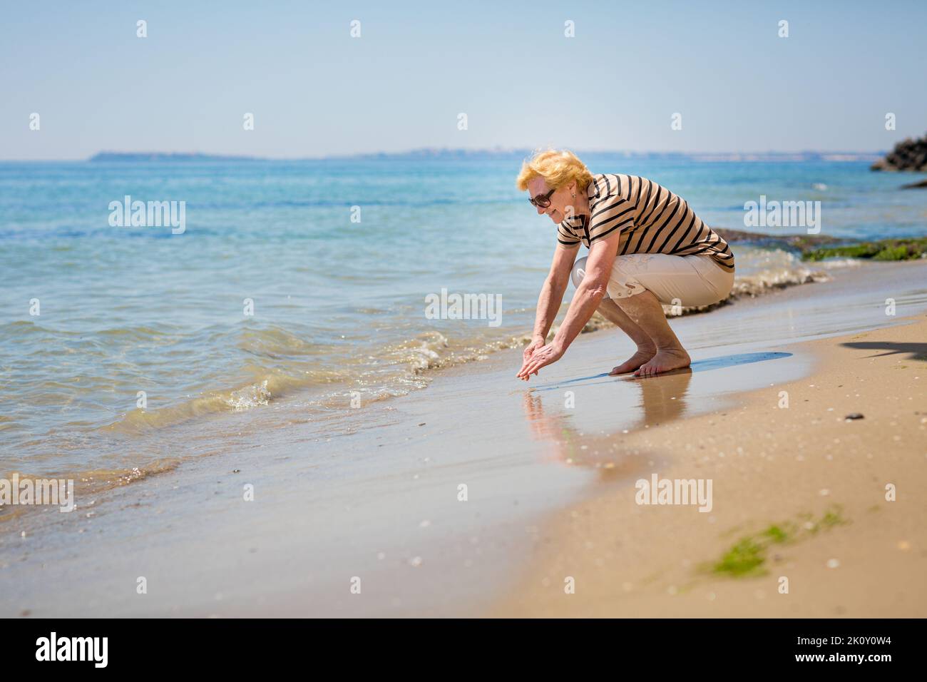 Mujer anciana atractiva tocando el agua en la orilla del mar Foto de stock