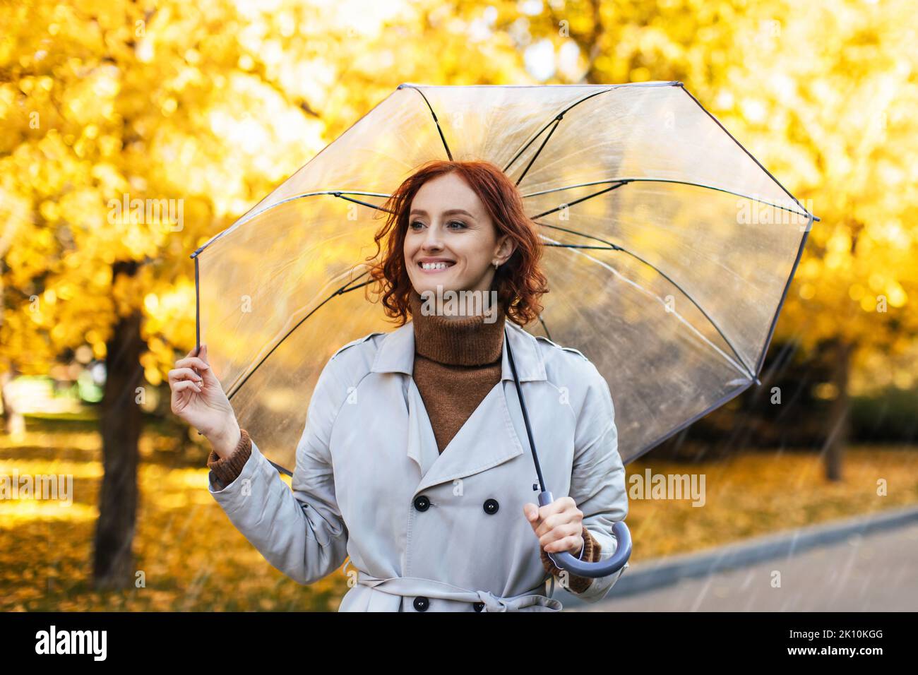 Alegre mujer caucásica milenaria de pelo rojo con chubasquero y paraguas disfruta de la lluvia en el parque Foto de stock