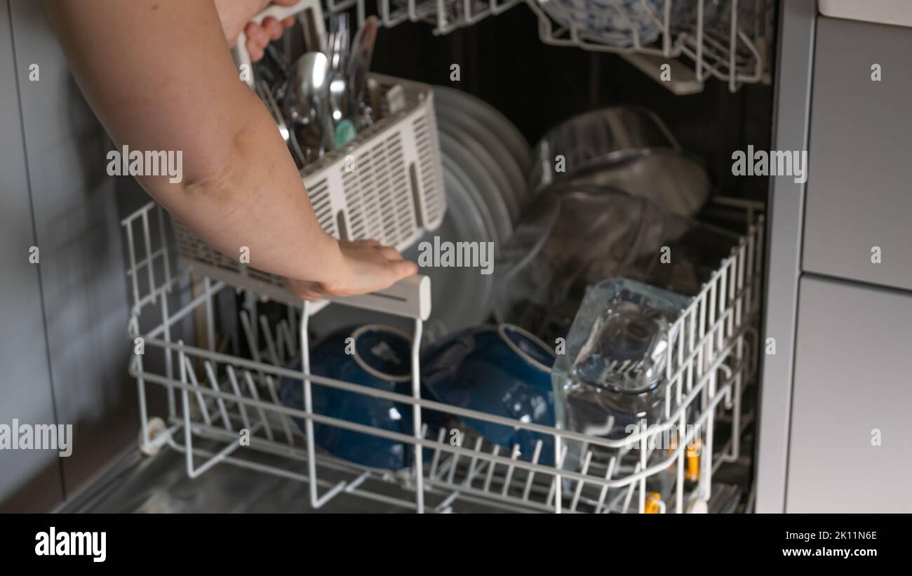 Desmontador del lavavajillas.Las manos de las mujeres clasifican los platos limpios del lavavajillas. Platos, tenedores y vasos en el lavavajillas.Lavavajillas con platos limpios. Foto de stock