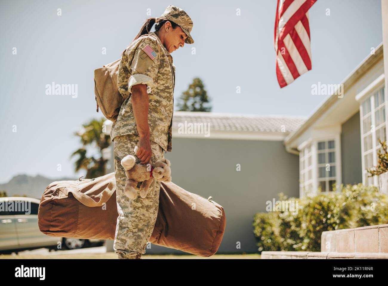 Madre militar estadounidense sosteniendo un osito de peluche mientras estaba de pie fuera de su casa con su equipaje. Soldado patriota volviendo a casa después de servir h Foto de stock