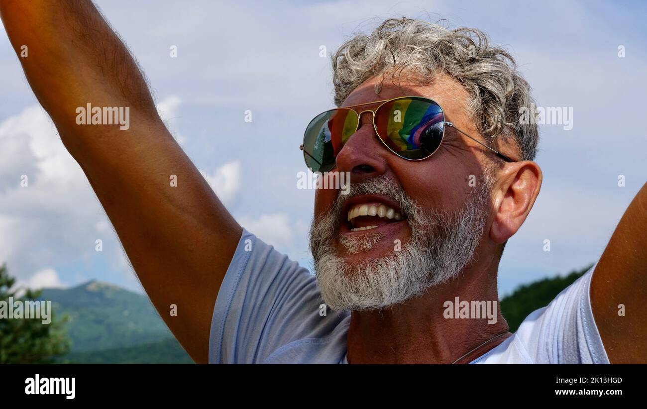 Retrato de un anciano caucásico de pelo gris con barba y gafas de sol sosteniendo una bandera LGBTQIA arco iris contra un fondo del cielo en desfile gay. Celebra el Mes del Orgullo, el Día de Salida Foto de stock