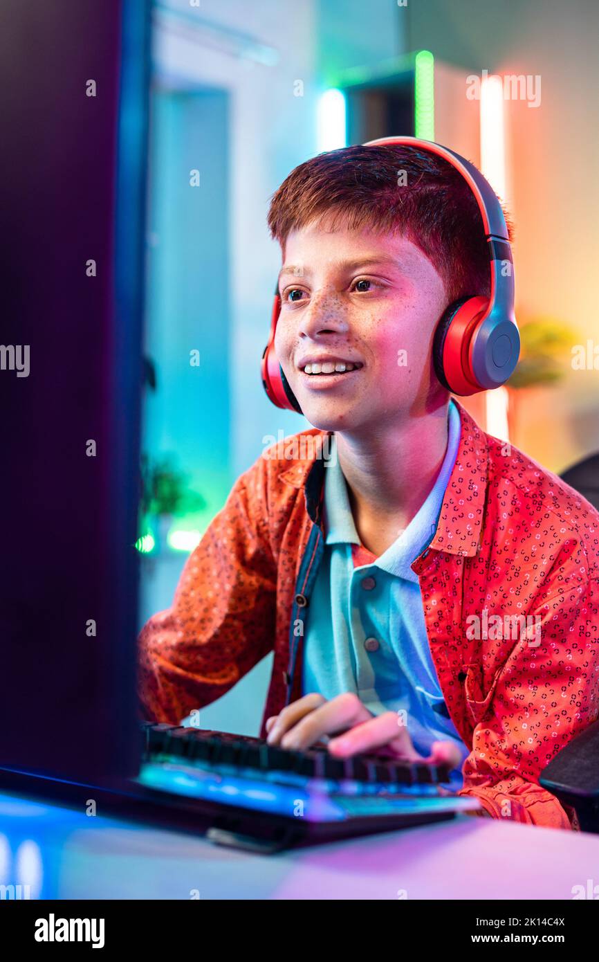 Toma vertical, niño adolescente feliz y sonriente que juega a videojuegos en directo en el ordenador hablando con auriculares inalámbricos en casa: Concepto de entretenimiento Foto de stock
