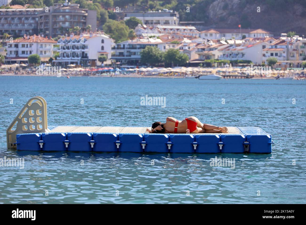 Mujer en bikini de color rojo tomar el sol mientras está acostada en pontón flotante en el agua del mar Mediterráneo en el fondo de la ciudad resort, vacaciones de playa Foto de stock