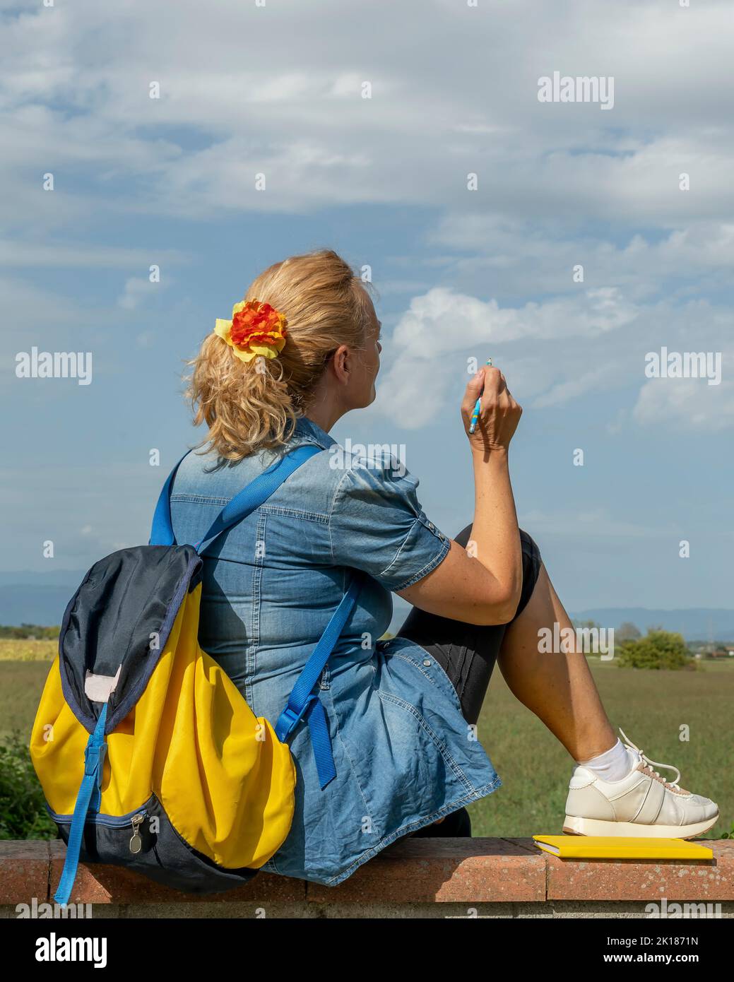 Mujer rubia con mochila sentada en una pared baja pretende dibujar en el cielo delante de ella Foto de stock