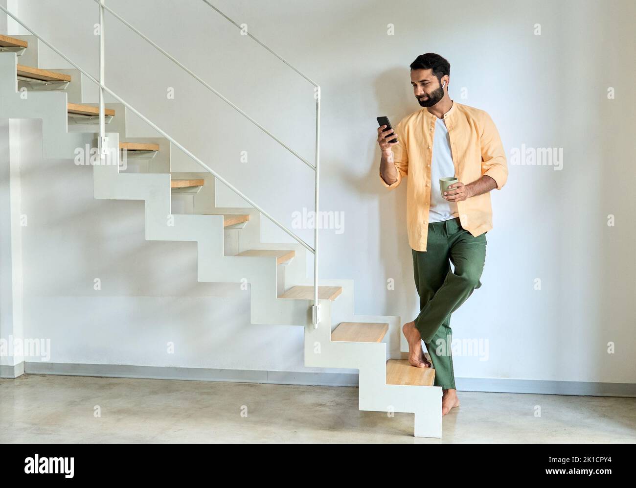 Un hombre indio que lleva un auricular con un smartphone de pie en casa. Foto de stock