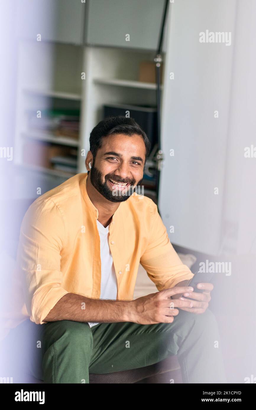 Hombre indio alegre sentado en casa usando el teléfono móvil escuchando música. Foto de stock