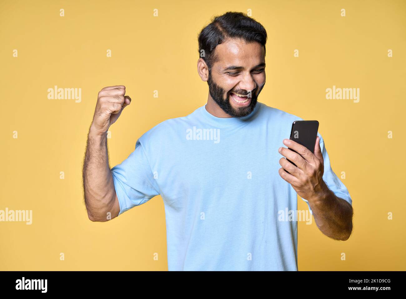 Hombre indio emocionado levantando el puño con el smartphone aislado sobre fondo amarillo. Foto de stock