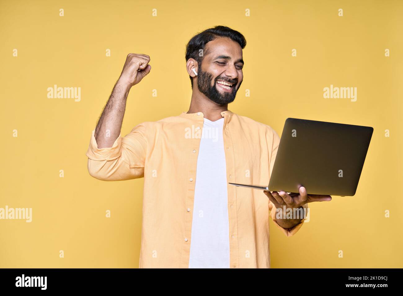 Hombre indio feliz levantando puño viendo el juego en el ordenador portátil aislado en amarillo. Foto de stock