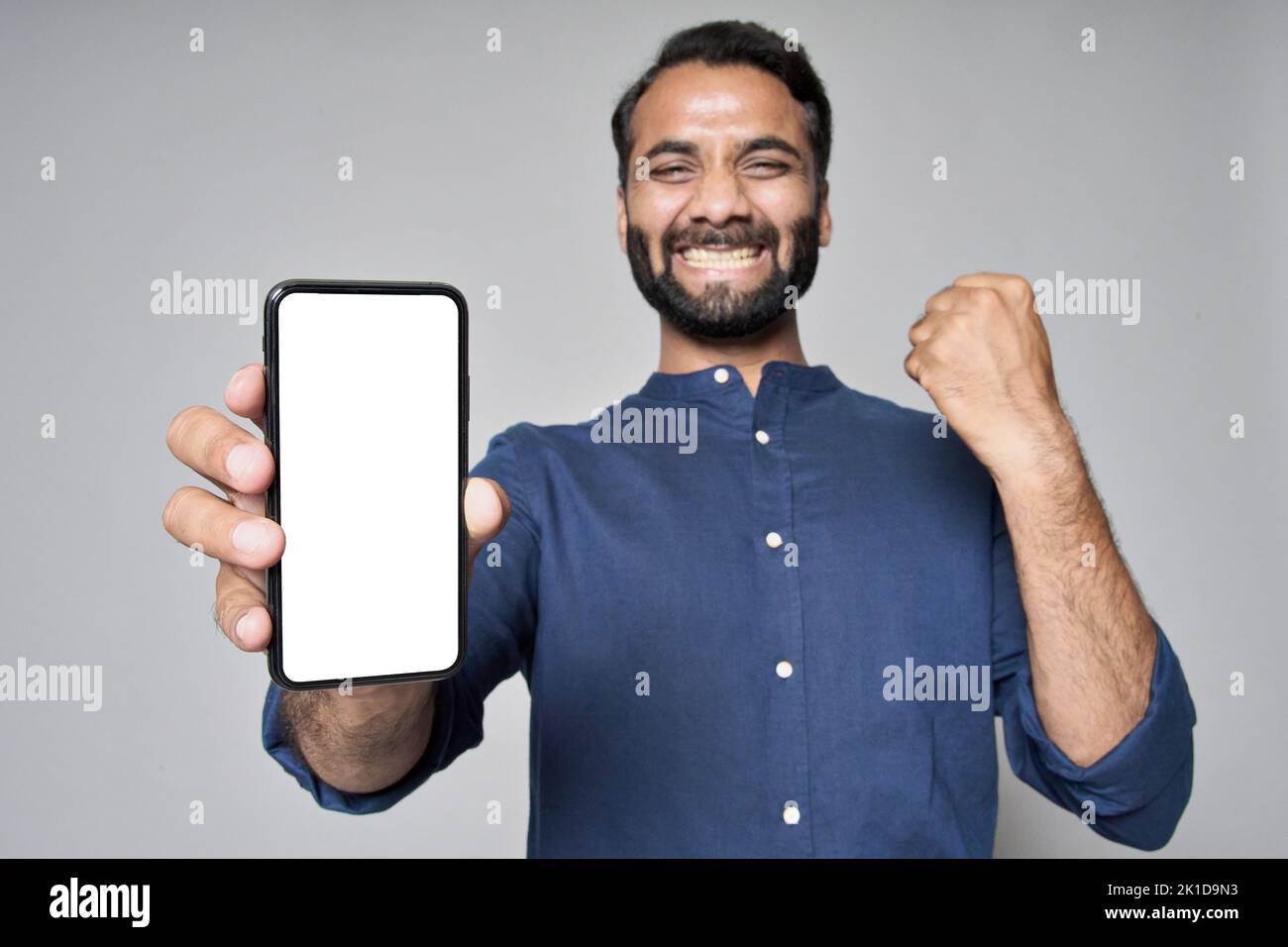 Feliz hombre de negocios indio aislado en gris mostrando teléfono móvil celebrando la victoria. Foto de stock