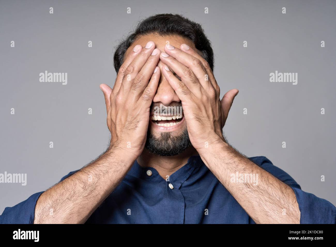 Hombre indio feliz cerrando los ojos con las manos aisladas sobre fondo gris. Foto de stock