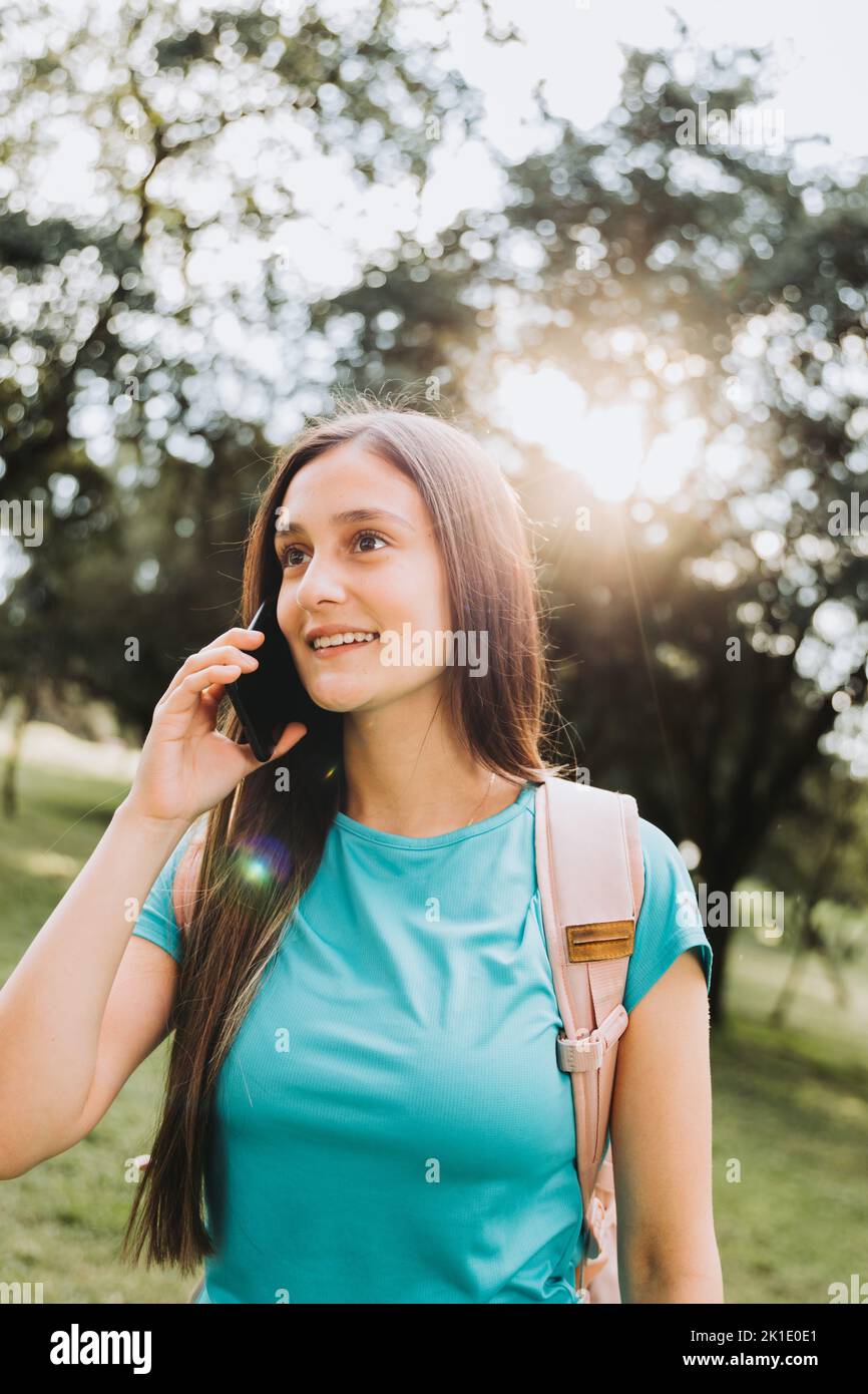 Adolescente niña estudiante con camisa de aguamarina, haciendo una llamada telefónica a su familia en el parque. Retroiluminación solar Foto de stock