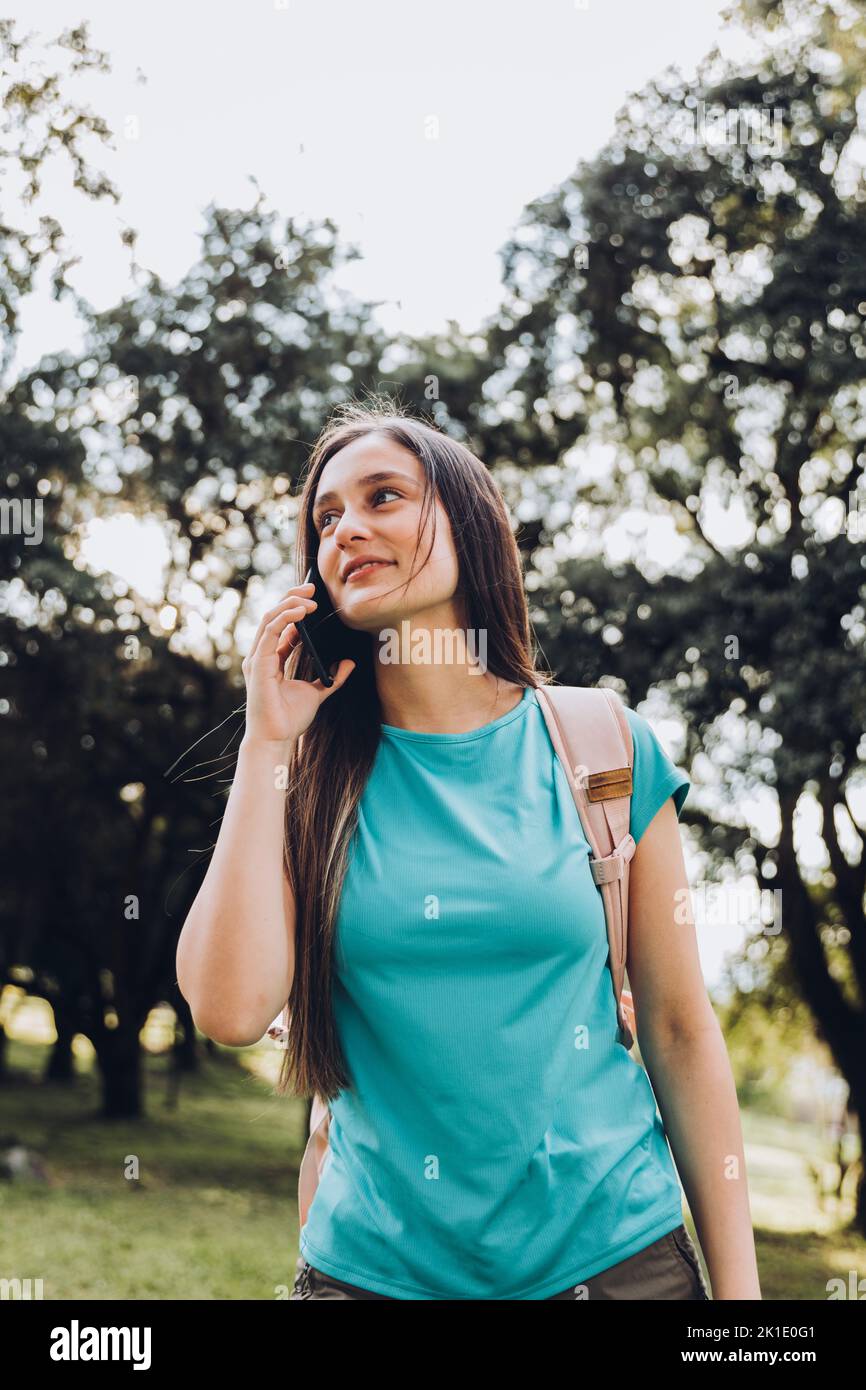 Adolescente niña estudiante con camisa de aguamarina, haciendo una llamada telefónica a su familia en el parque. Retroiluminación solar Foto de stock