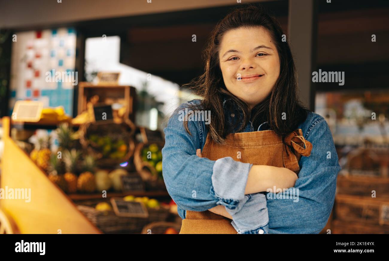 Empleado de la tienda con síndrome de Down sonriendo a la cámara mientras está de pie en una tienda de comestibles. Mujer empoderada con una discapacidad intelectual que trabaja en a. Foto de stock