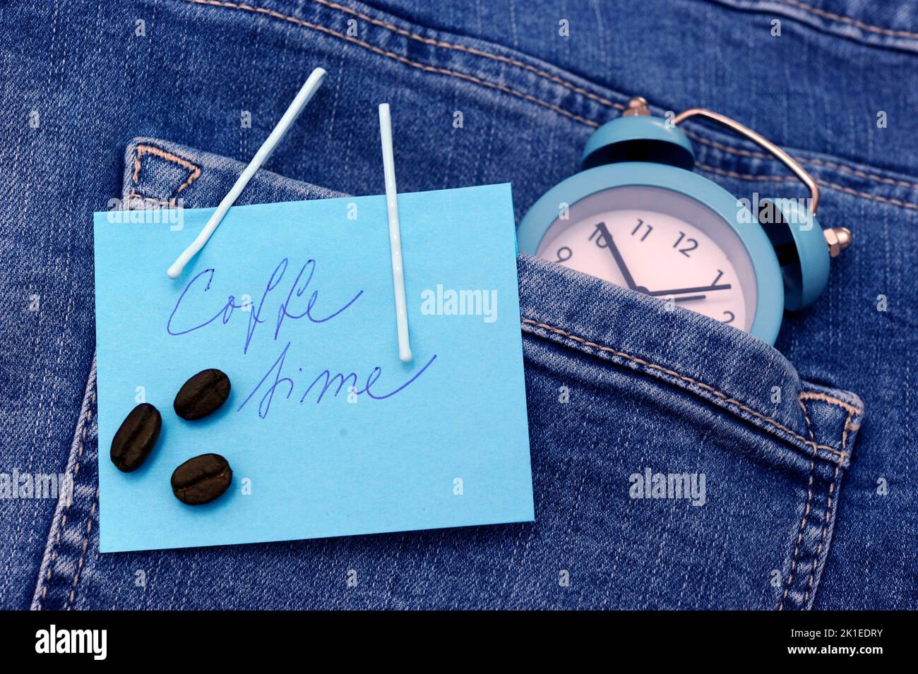 reloj despertador azul con granos de café taza de café expreso sobre jeans o vaqueros de bolsillo como el concepto de tiempo y el amor por el café o el café de la mañana o el café t Foto de stock