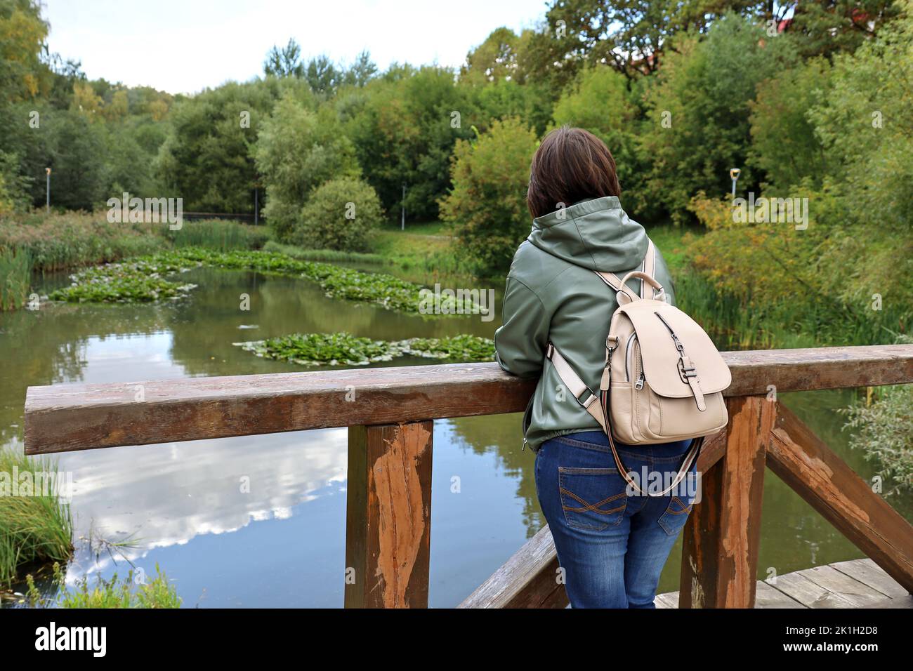 Mujer solitaria de pie sobre un puente de madera sobre un pintoresco fondo de estanque. Parque de otoño, ocio en la naturaleza Foto de stock