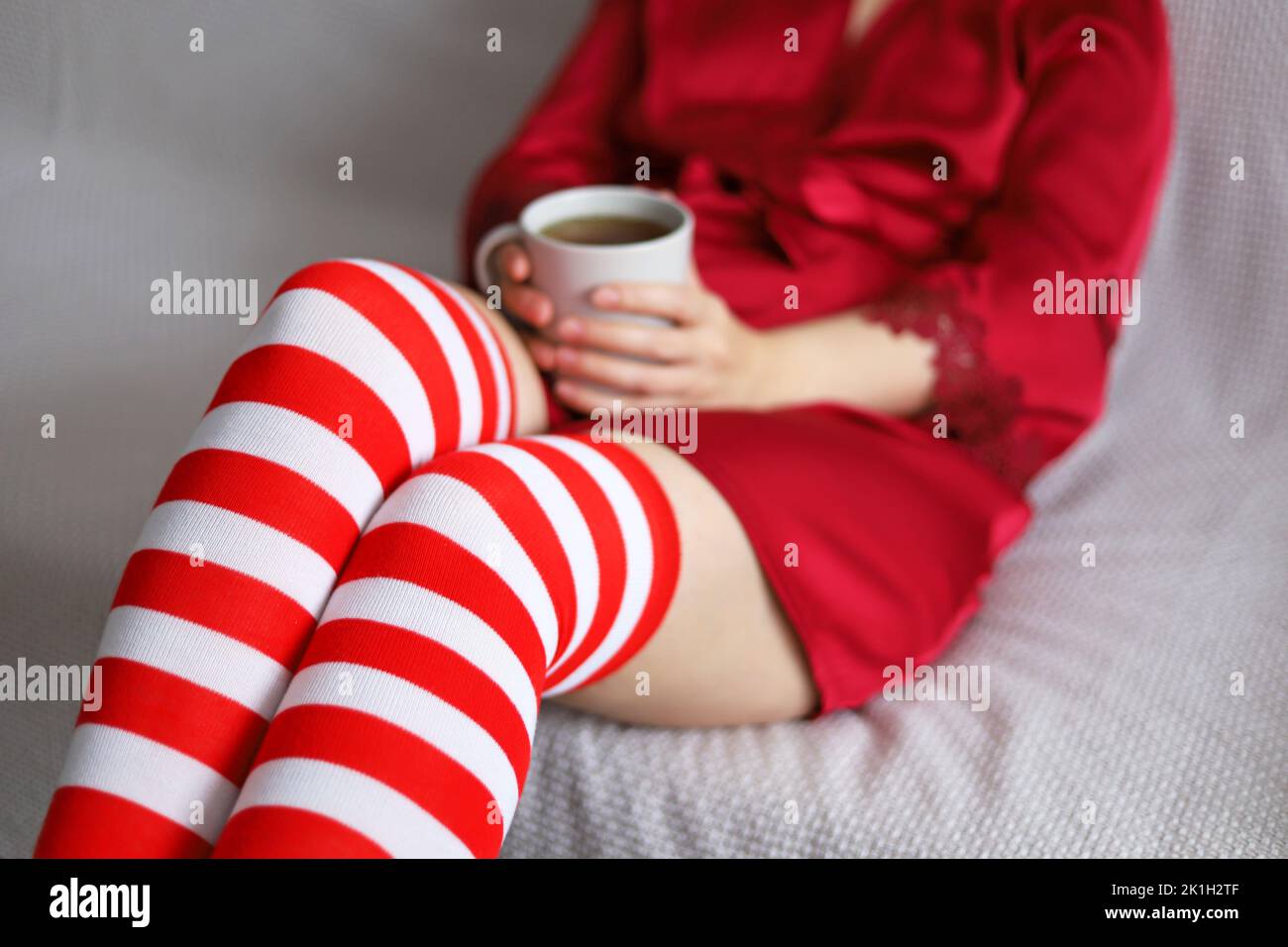 Mujer con calcetines de rodilla de color rojo blanco y camisón de seda sentada en un sofá con taza de té en las manos. Vacaciones de Navidad en casa, bebida caliente en invierno Foto de stock