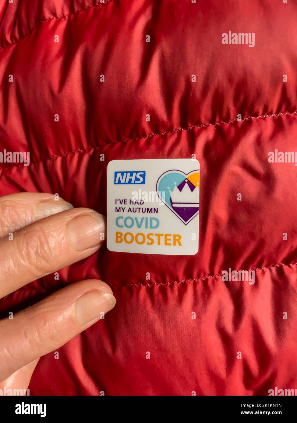 Hampshire, Inglaterra, Reino Unido. 2022. Pegatina adhesiva ofrecida por la NHS para tener un otoño covid refuerzo jab en una chaqueta acolchada roja. Foto de stock