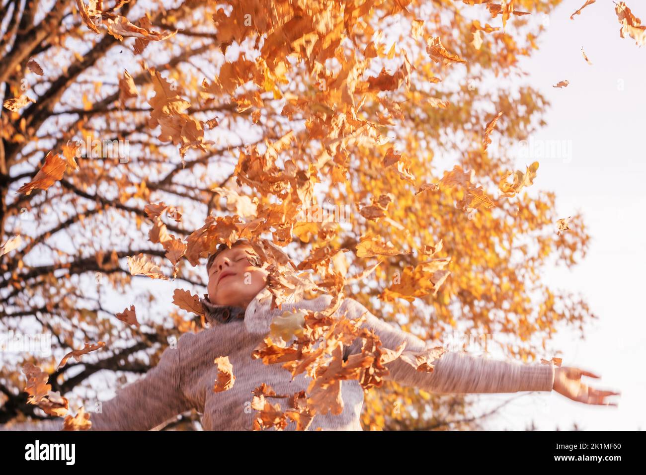 Niño de 9-10 años de edad se encuentra en el parque de otoño en rayos de puesta de sol y lanza hojas de roble, sonriendo y cerrando sus ojos. Niño desenfocado en gris Foto de stock