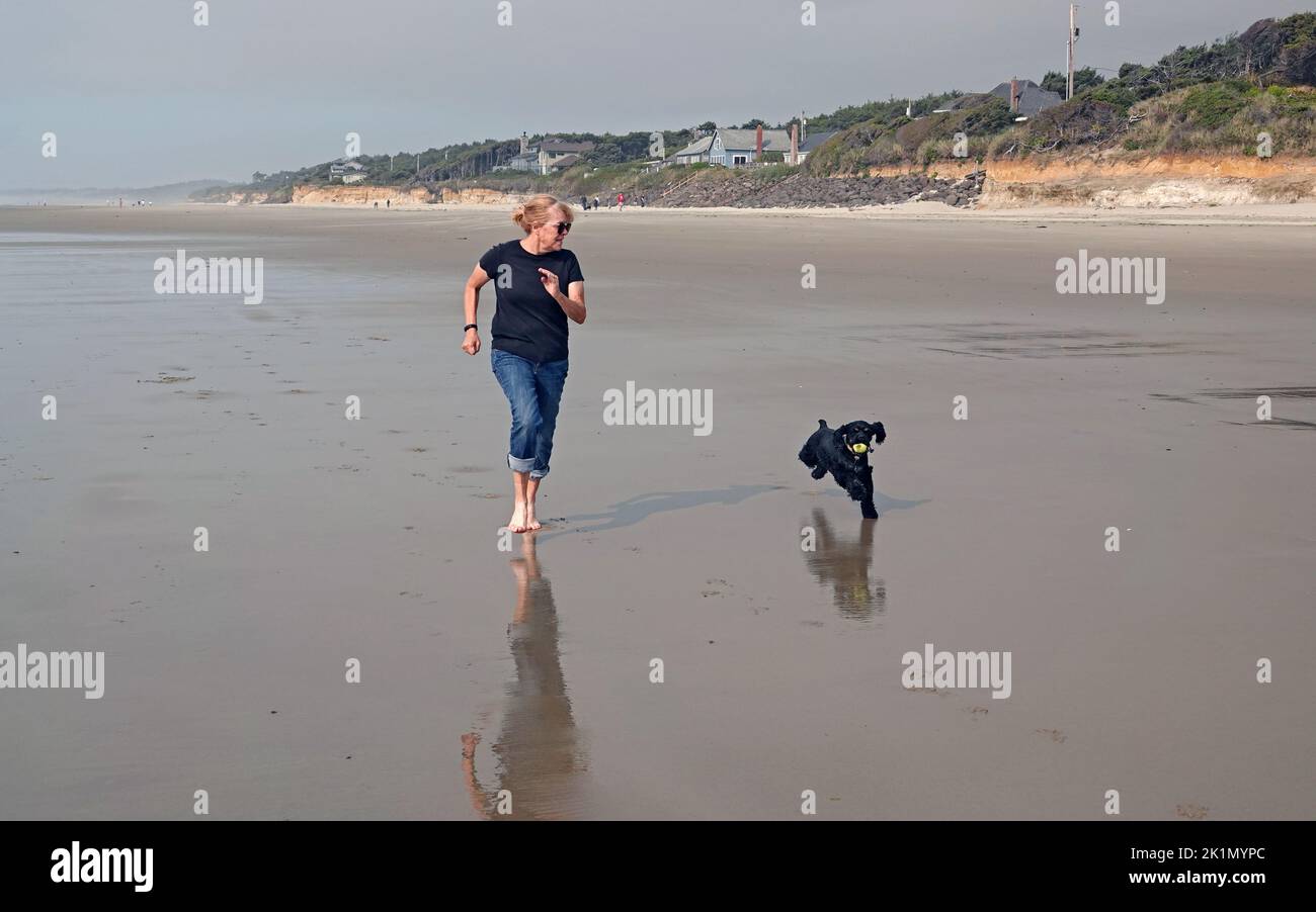 Una mujer mayor corre a lo largo de una playa cerca de Yachats, Oregon, con su cachorro spaniel. Foto de stock