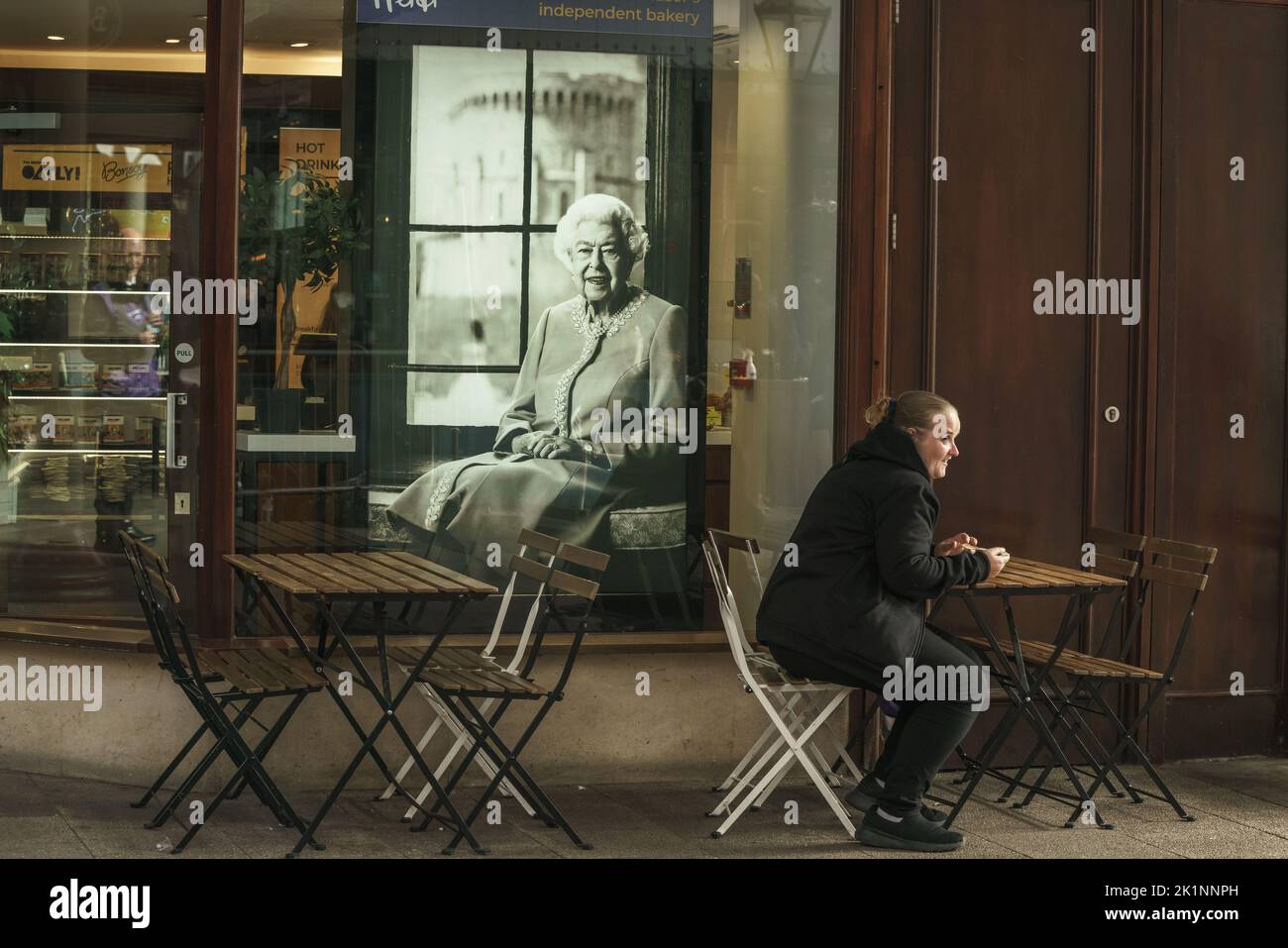 Una mujer se sienta sola en una mesa en un café de Windsor, a la que se ve un retrato de Su Majestad la Reina Isabel II Té con la Reina. Foto de stock