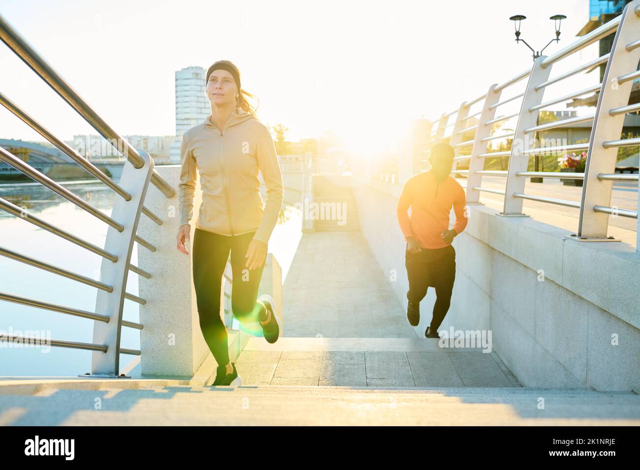 Vista frontal de la joven deportista corriendo a lo largo de un puente moderno con un hombre afroamericano en activewear sobre el fondo Foto de stock