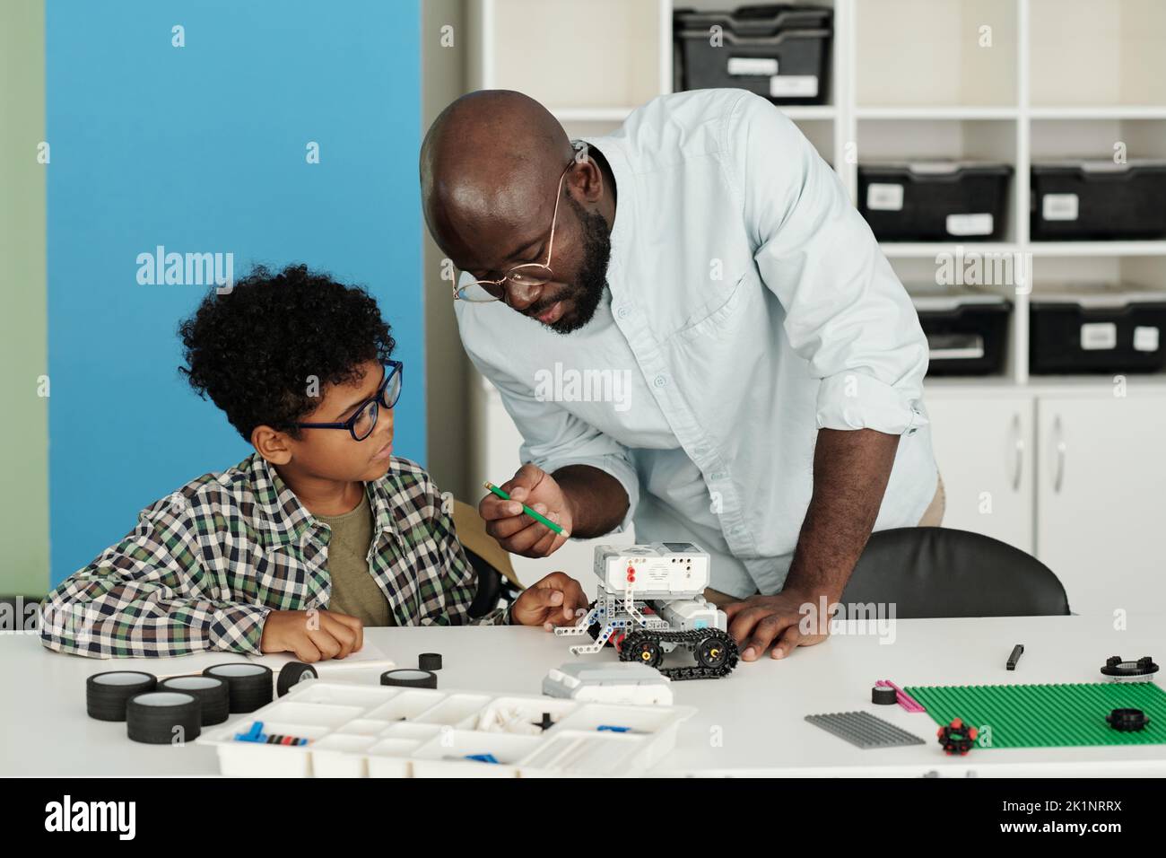 Joven maestro afroamericano de robótica señalando a juguete mientras explica cómo conectar partes de nuevo modelo de robot Foto de stock