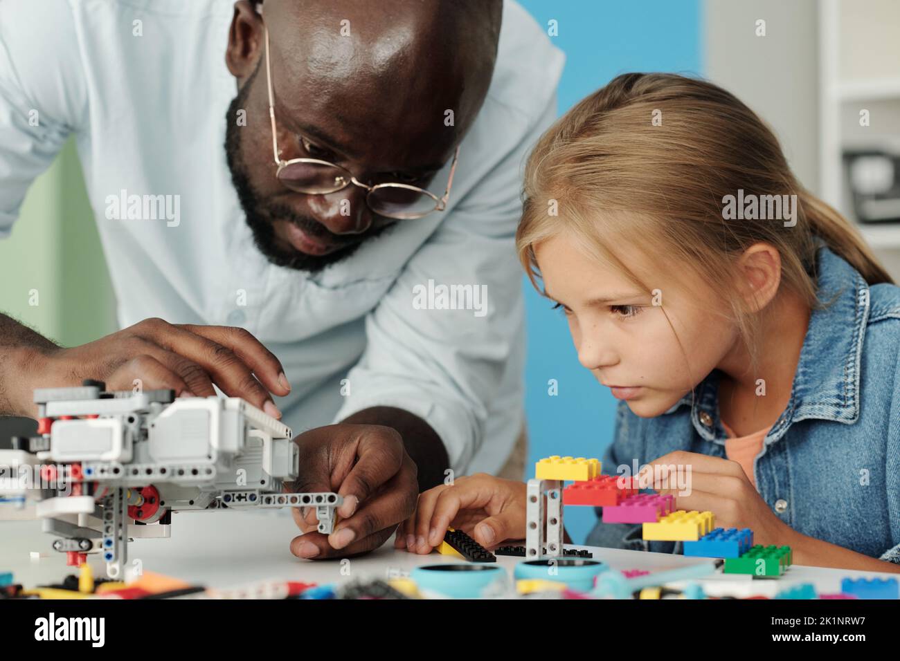 Joven afroamericano inclinándose sobre el escritorio mientras ayudaba a la alumna con la construcción de un nuevo robot en la lección de robótica en la escuela Foto de stock
