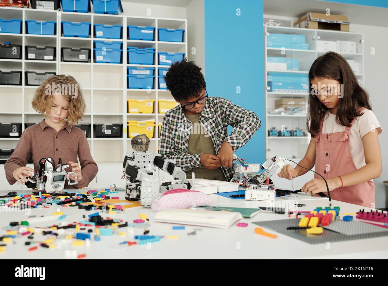 Tres estudiantes inteligentes de la escuela primaria construyendo nuevos robots en la lección mientras que de pie junto a la mesa con partes para conectar juntos Foto de stock