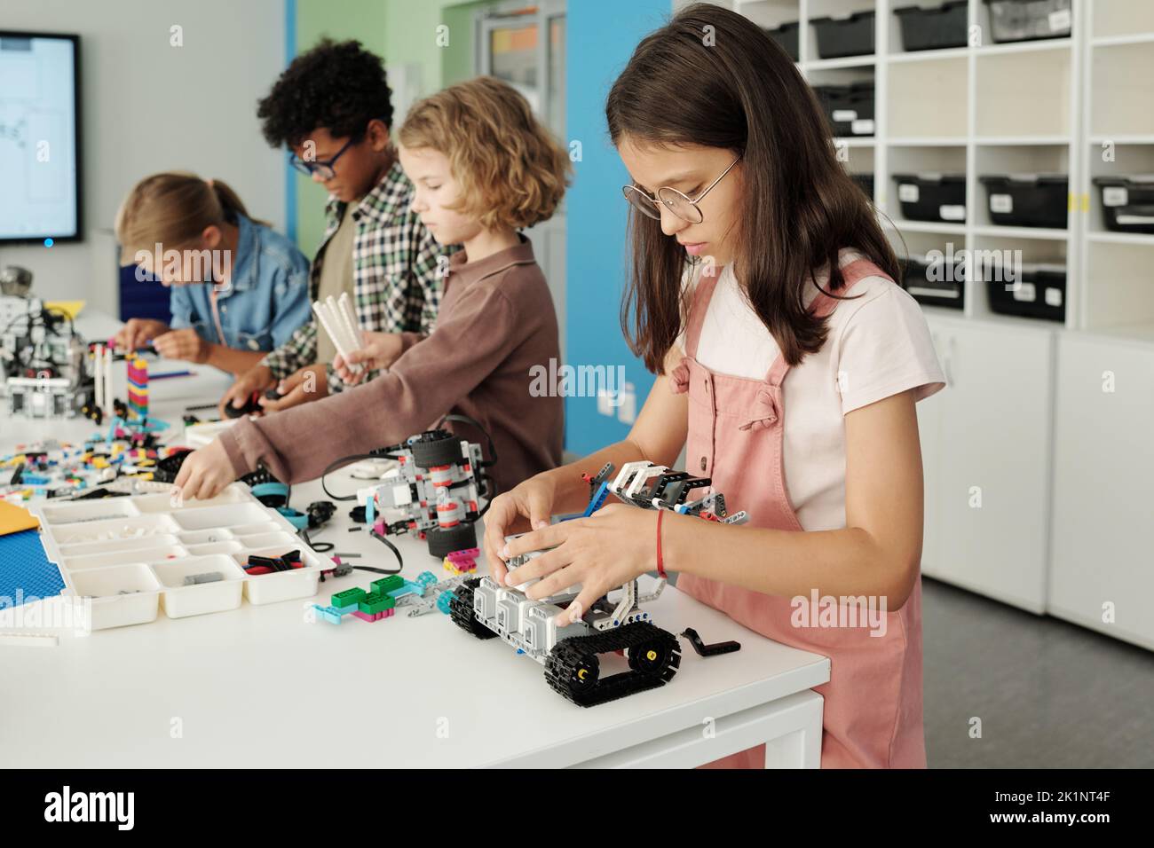 Fila de cuatro estudiantes interculturales de la escuela primaria construyendo nuevos robots de juguete mientras que de pie por la mesa con detalles en el aula Foto de stock