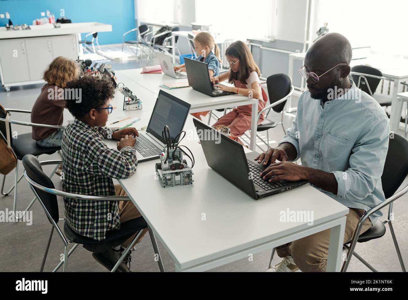 Grupo de alumnos interculturales creativos y su maestro usando computadoras portátiles en la lección de robótica mientras se sientan uno frente al otro Foto de stock