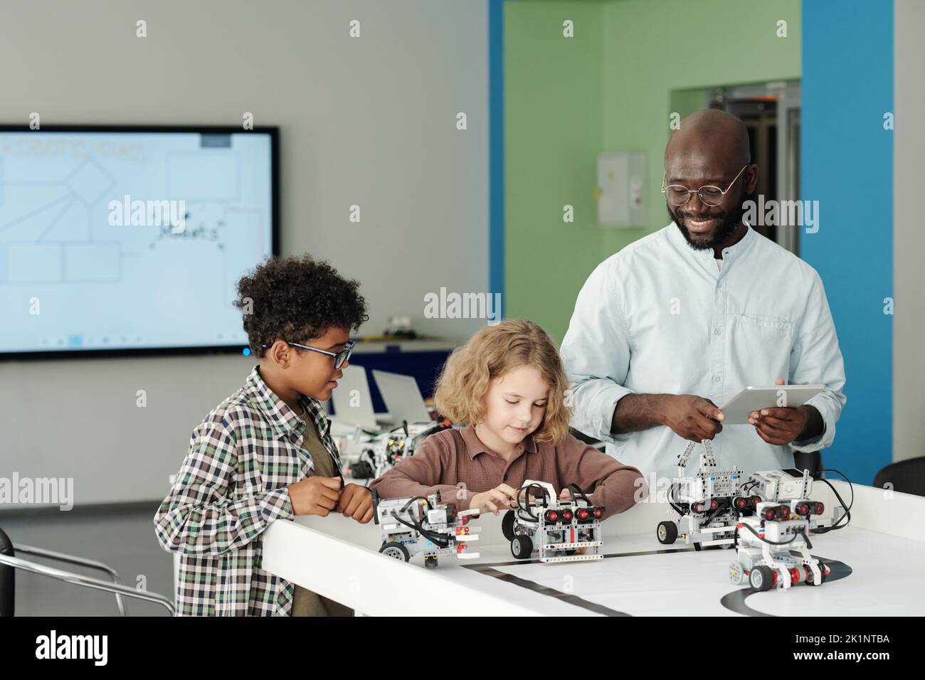 Feliz joven profesor de robótica con tableta de pie por dos inteligentes escolares interculturales jugando con nuevos modelos de robots Foto de stock
