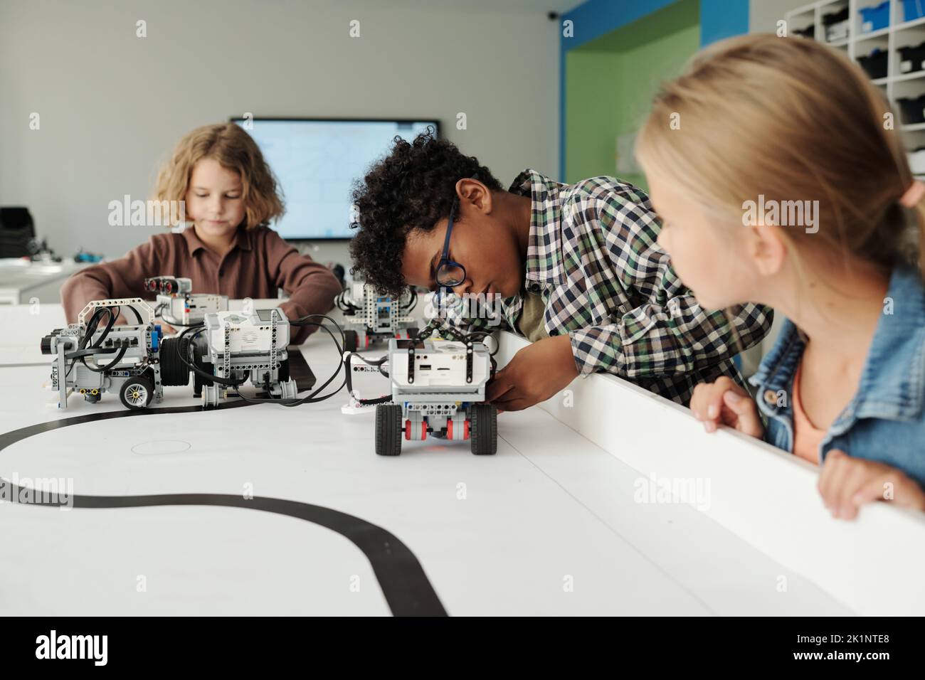Grupo de escolares interculturales lindos jugando con nuevos modelos de robots a la mesa mientras los niños controlando el movimiento de los juguetes Foto de stock