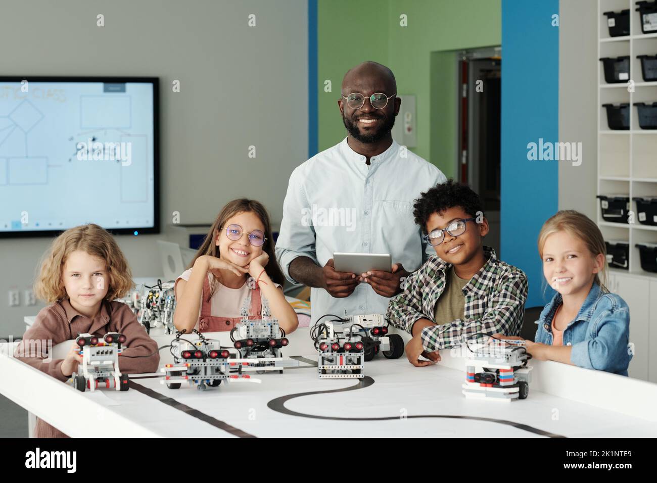 Grupo de jóvenes y jóvenes de escuela intercultural feliz y su maestro mirando la cámara mientras juegan con los nuevos modelos de robots en la lección Foto de stock